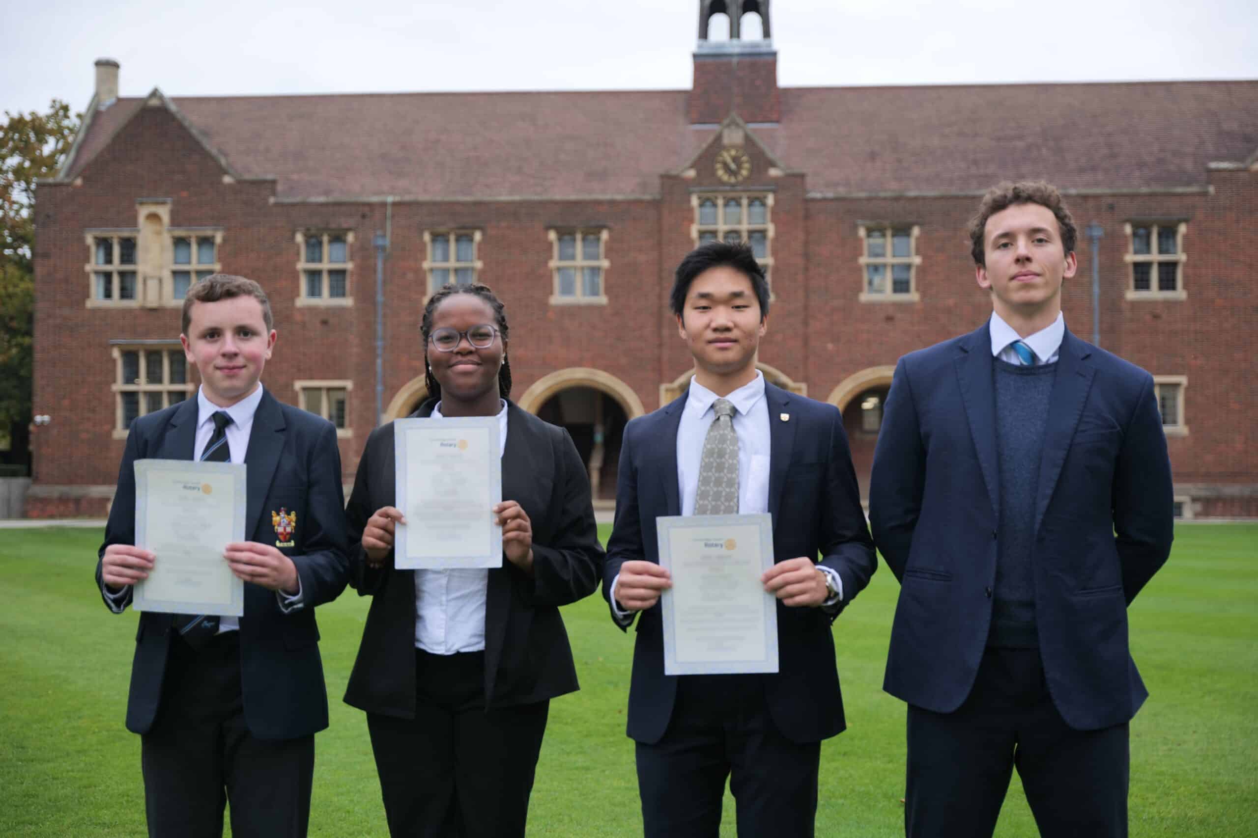 4 pupils stand in a row, the school behind them. They present their certificates.